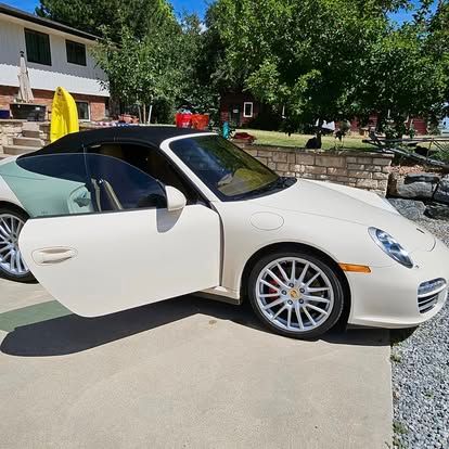 A cream-colored Porsche convertible with its driver-side door open, parked on a concrete driveway in front of a house.