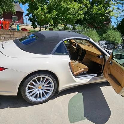 A cream-colored Porsche convertible with a black soft top and tan interior, parked on a concrete surface.