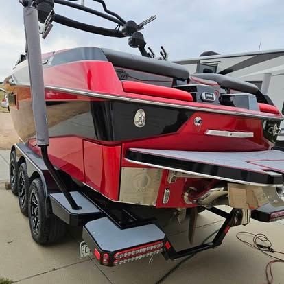 A rear view of a red and black wakeboard boat on a trailer, featuring a swim platform and a metal tower.