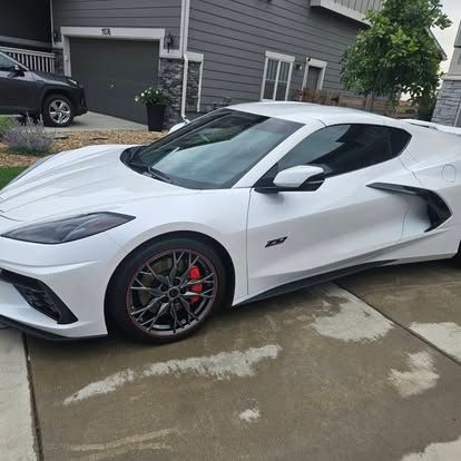 A white Chevrolet Corvette C8 parked on a residential driveway in front of a gray house.