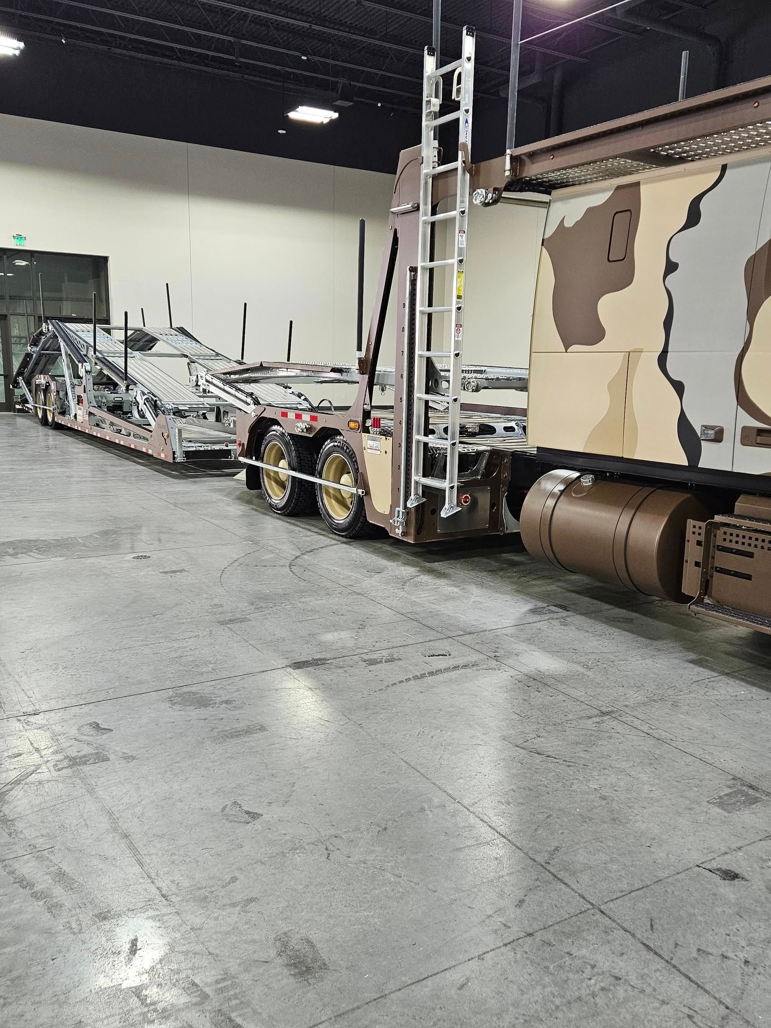 A desert-camouflage truck with an attached flatbed trailer parked inside a large, industrial warehouse.