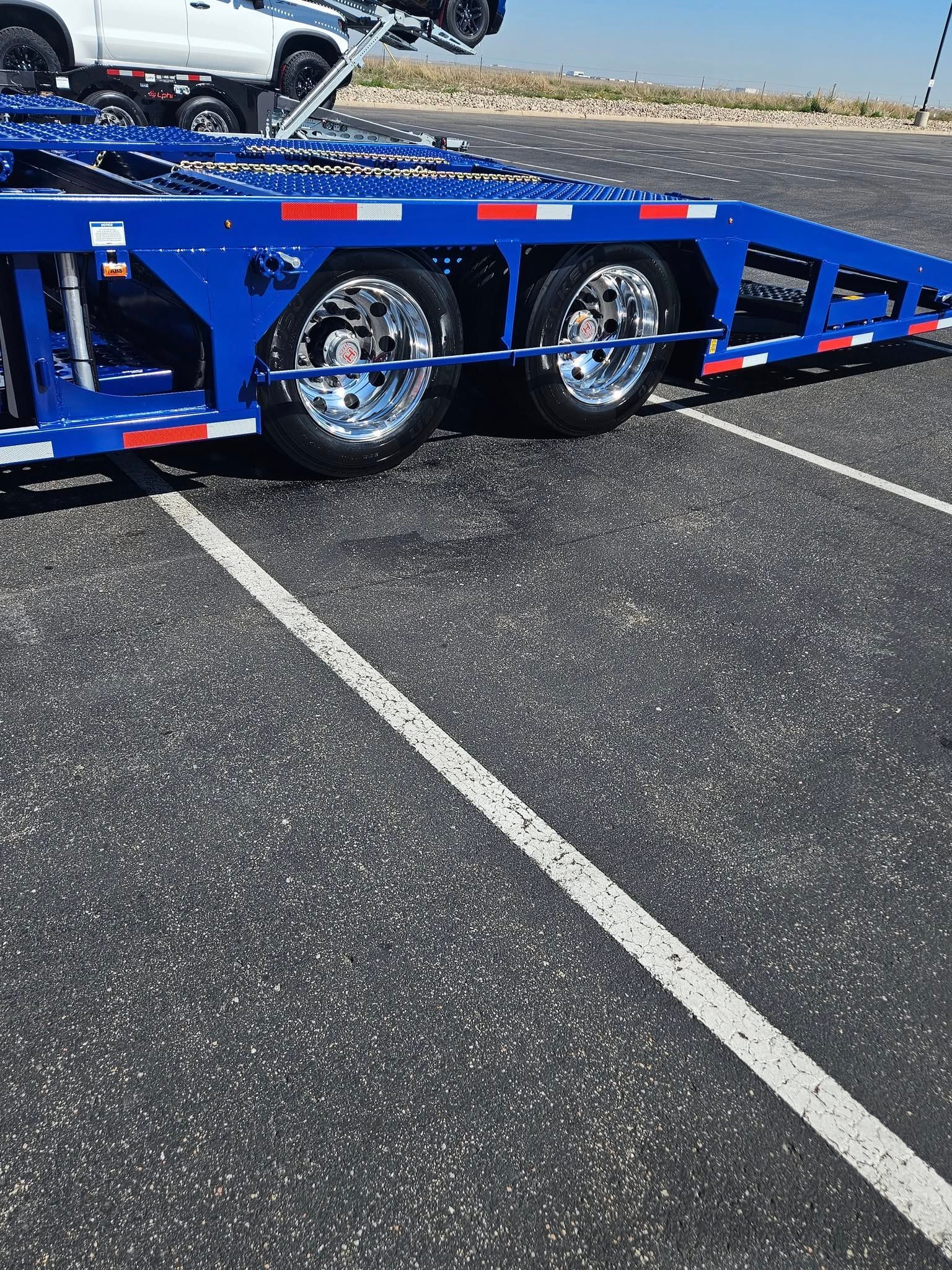 A close-up view of a blue car hauler trailer on a paved lot, featuring two chrome wheels and red and white reflectors.