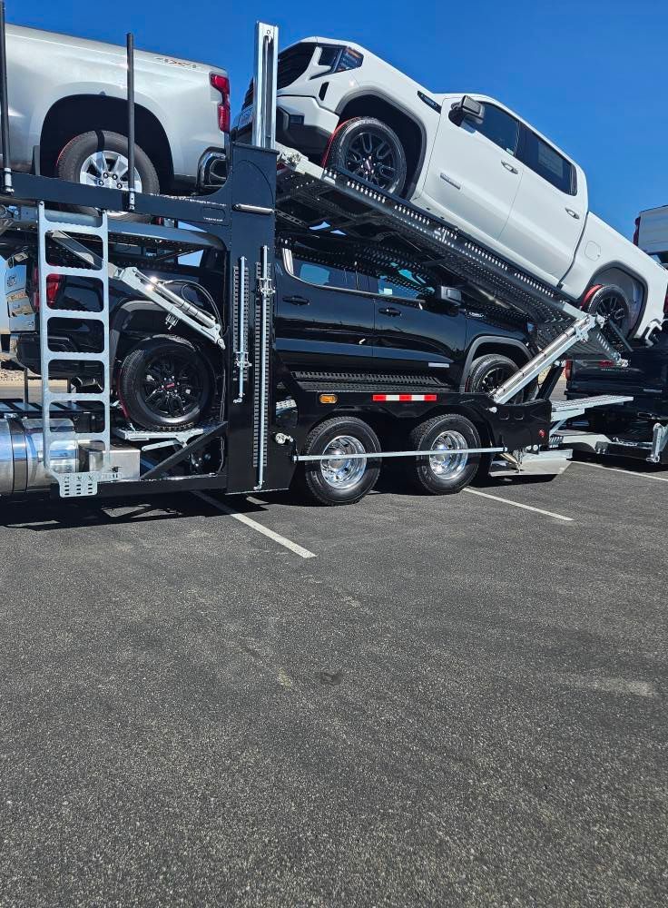 A car hauler trailer parked on asphalt, carrying three pickup trucks—two white and one black—on its upper and lower decks.