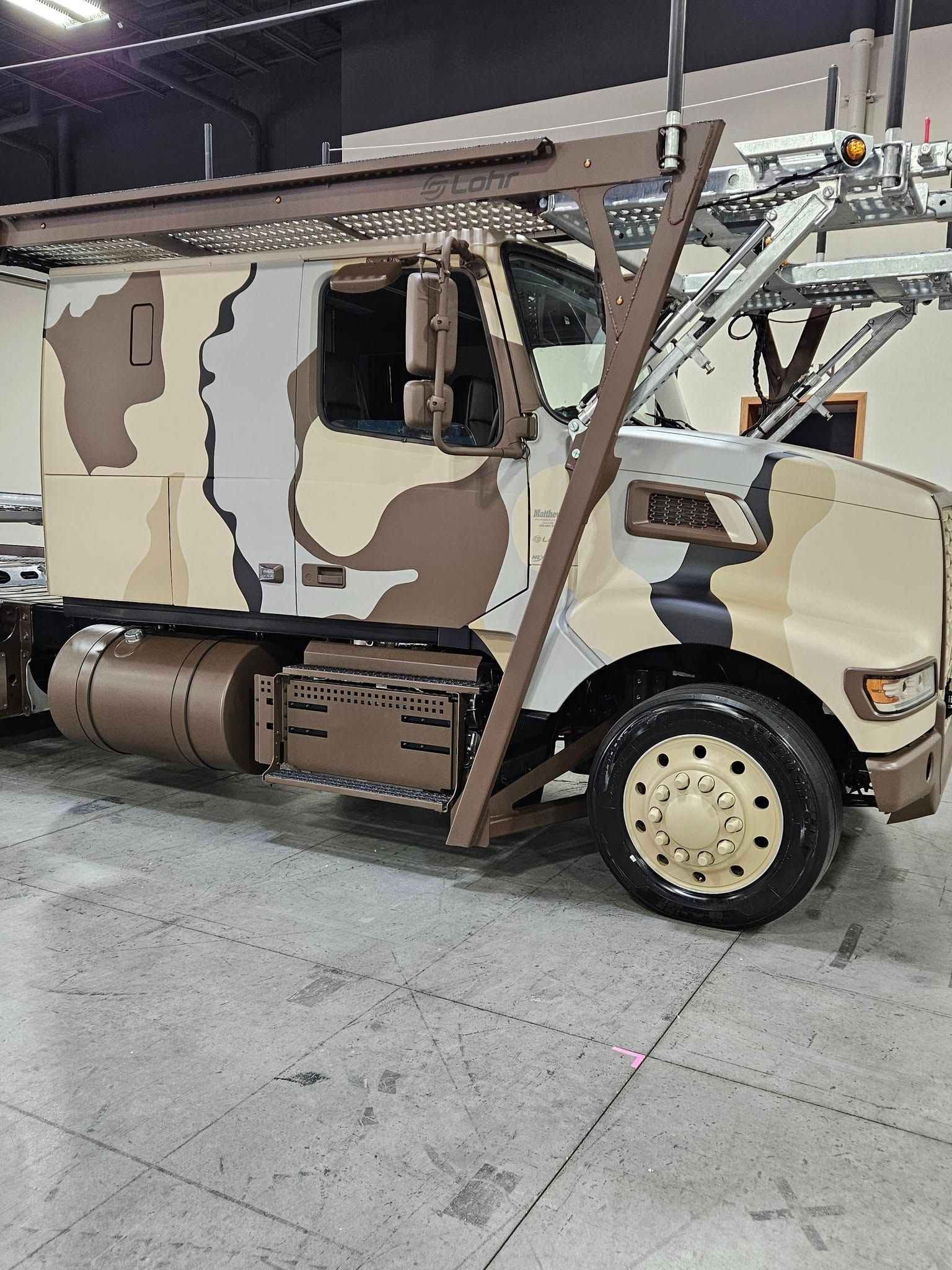 A desert camouflage-patterned semi-truck with a car hauler rack, parked inside a large, industrial-style building.