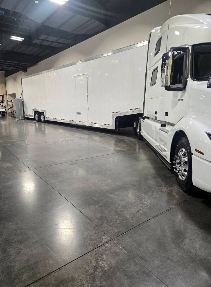A large white semi-truck with a long, white enclosed trailer parked on a concrete floor inside a warehouse.