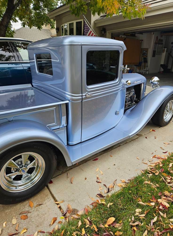 A light blue, vintage-style custom hot rod truck parked on a driveway with fallen autumn leaves.