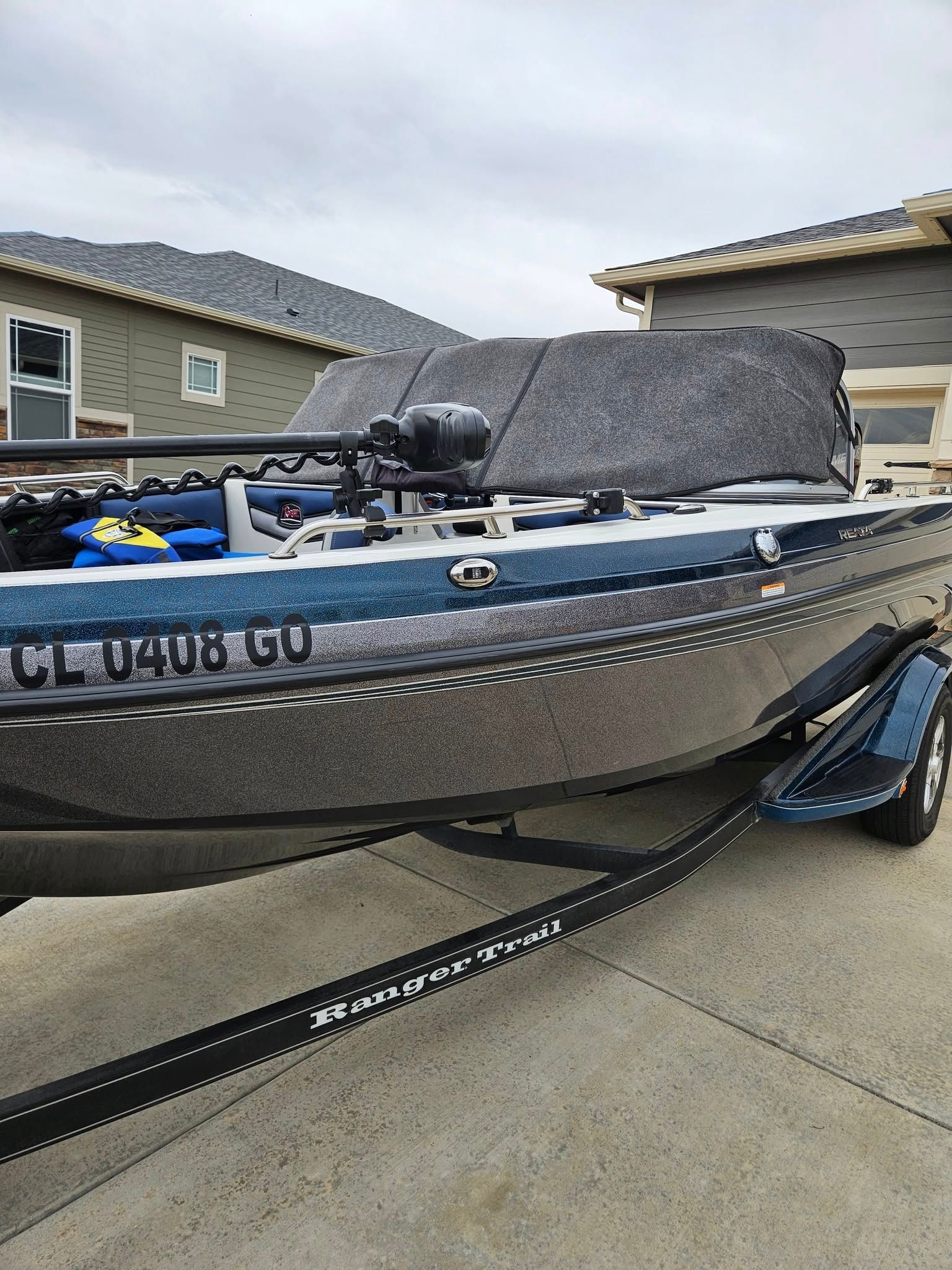 A bass fishing boat with a gray protective cover, hitched to a trailer on a paved driveway in front of suburban houses.