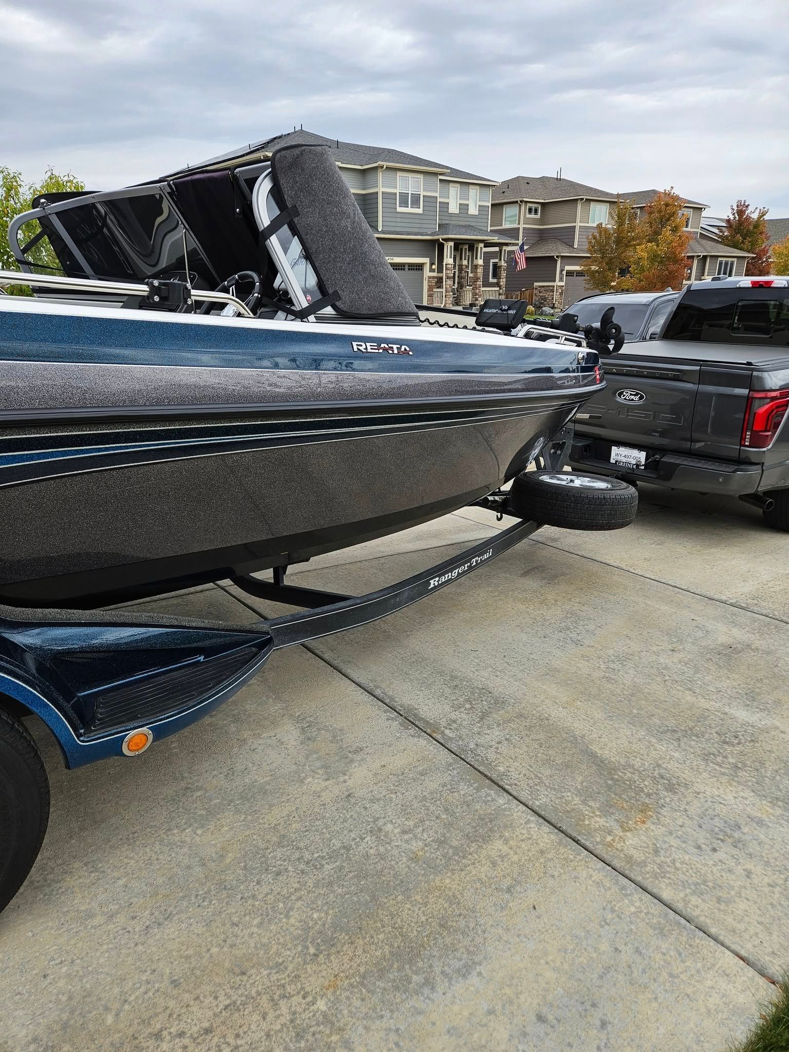 A blue fishing boat on a trailer parked in a driveway next to a grey pickup truck.