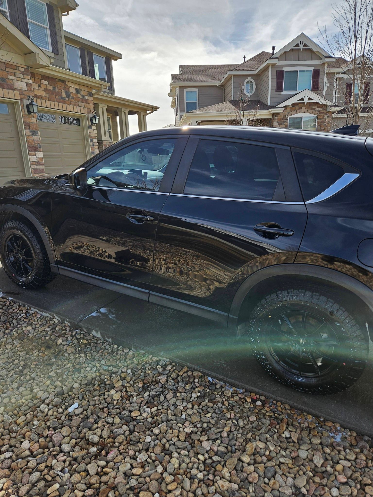 A black SUV parked on a pebble driveway in front of suburban houses under a partly cloudy sky.