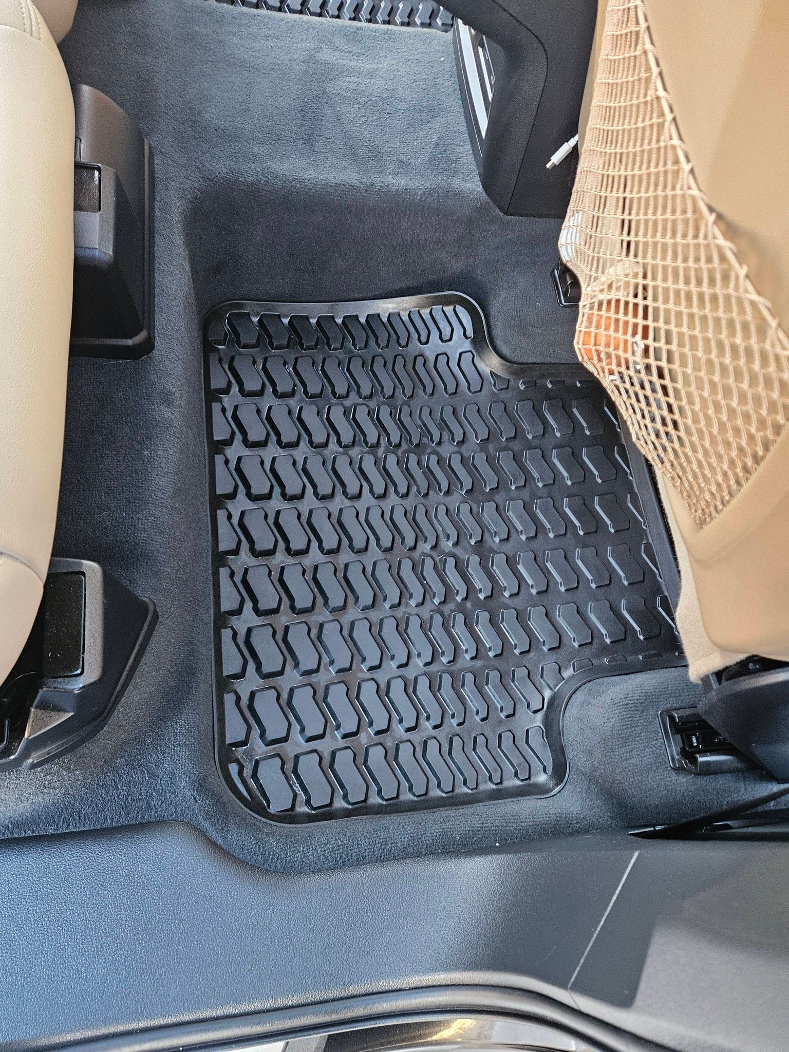 A black, textured rubber floor mat installed in the front footwell of a car with tan interior trim.