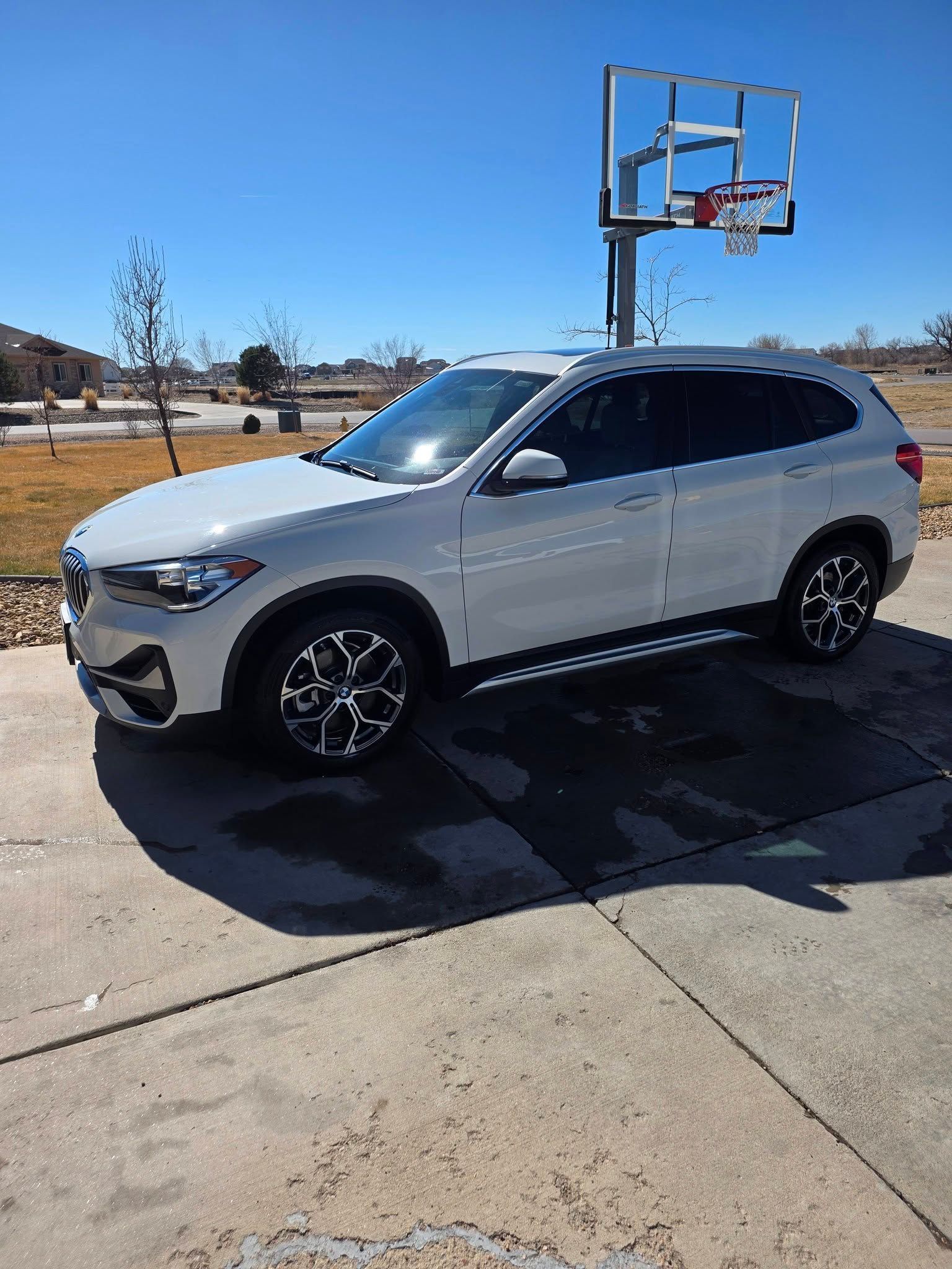 A white BMW SUV parked on a concrete driveway next to a basketball hoop on a sunny day.