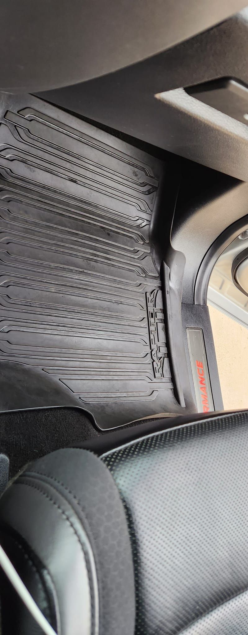 A close-up, high-angle view of a black car floor mat with textured, patterned rubber, situated by a leather vehicle seat.
