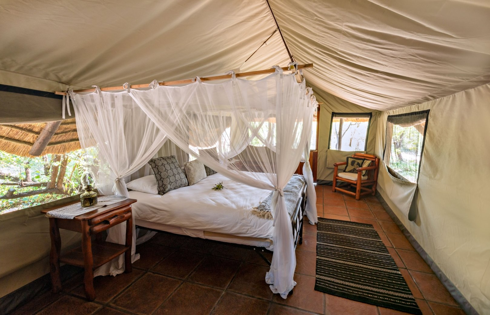 A safari-style tent interior featuring a canopy bed with white mosquito netting, a wooden bedside table, and an armchair.