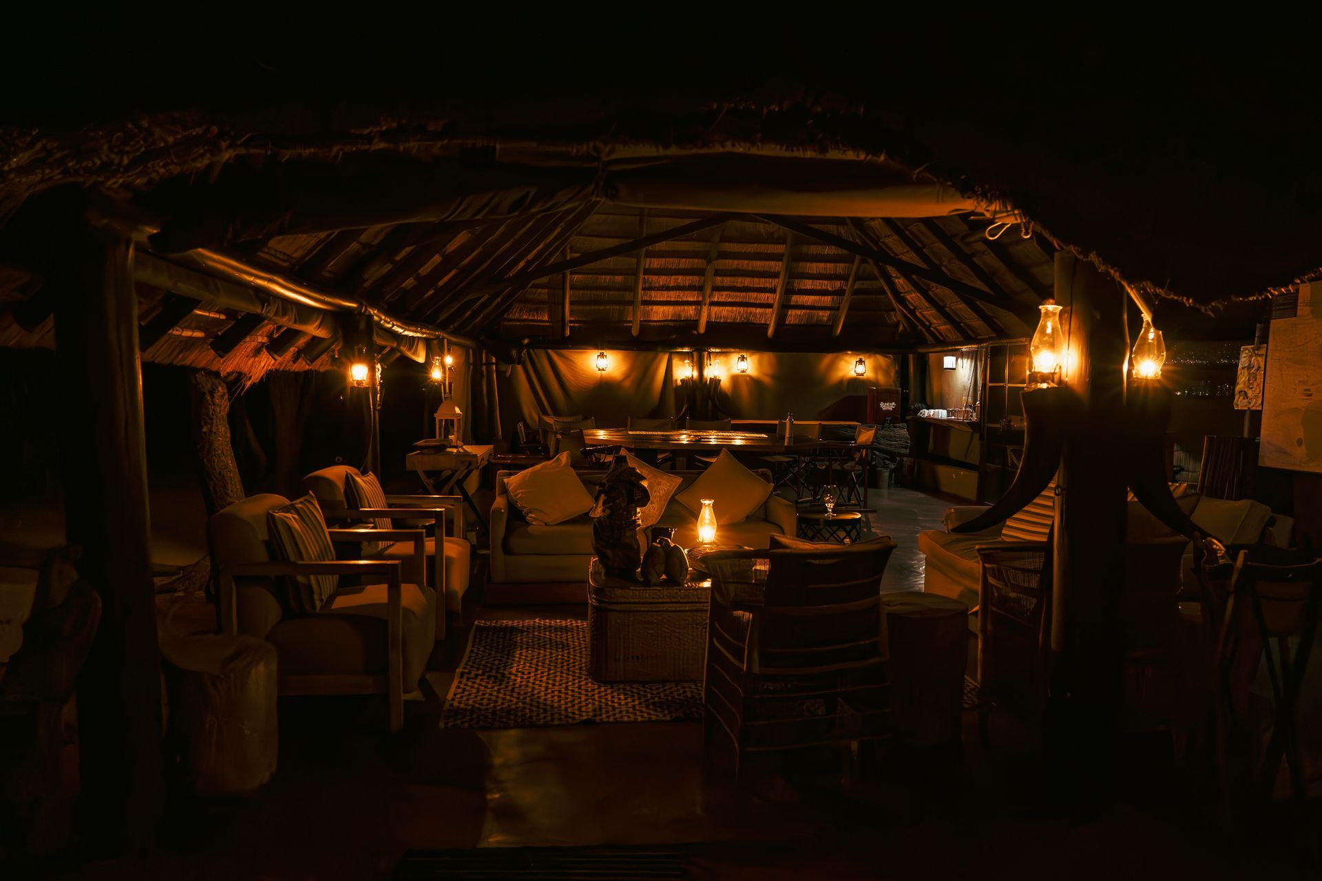 A rustic open-air lounge at night, featuring dimly lit lanterns, comfortable seating, and a thatched roof structure.