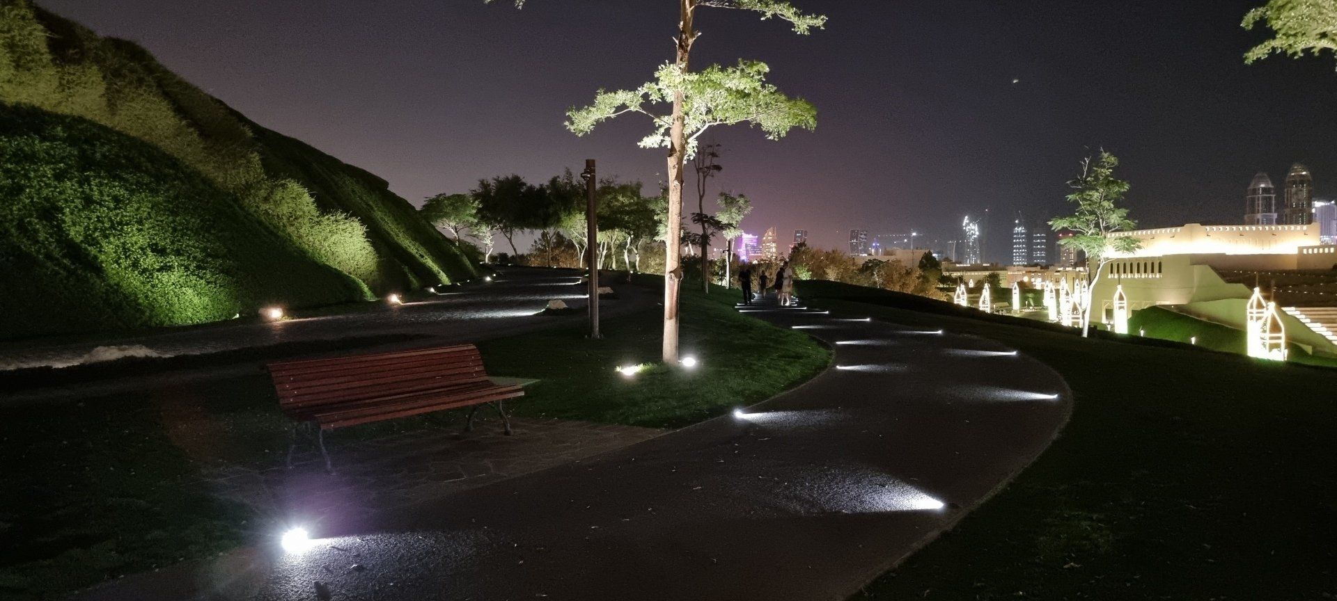 Curved pathway with modern lighting and a bench at Katara Hills, with city lights in the background.