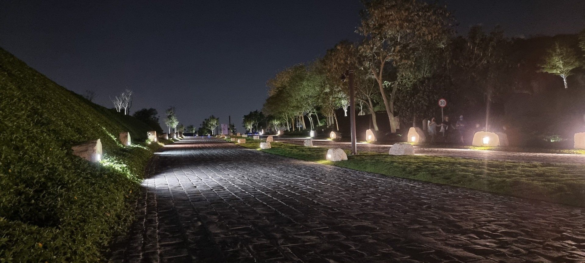 Cobblestone pathway at Katara Hills illuminated by modern lights, surrounded by trees and greenery.
