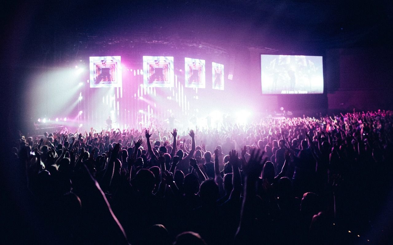 Silhouetted crowd with raised hands faces a brightly lit stage with performing band and screens.