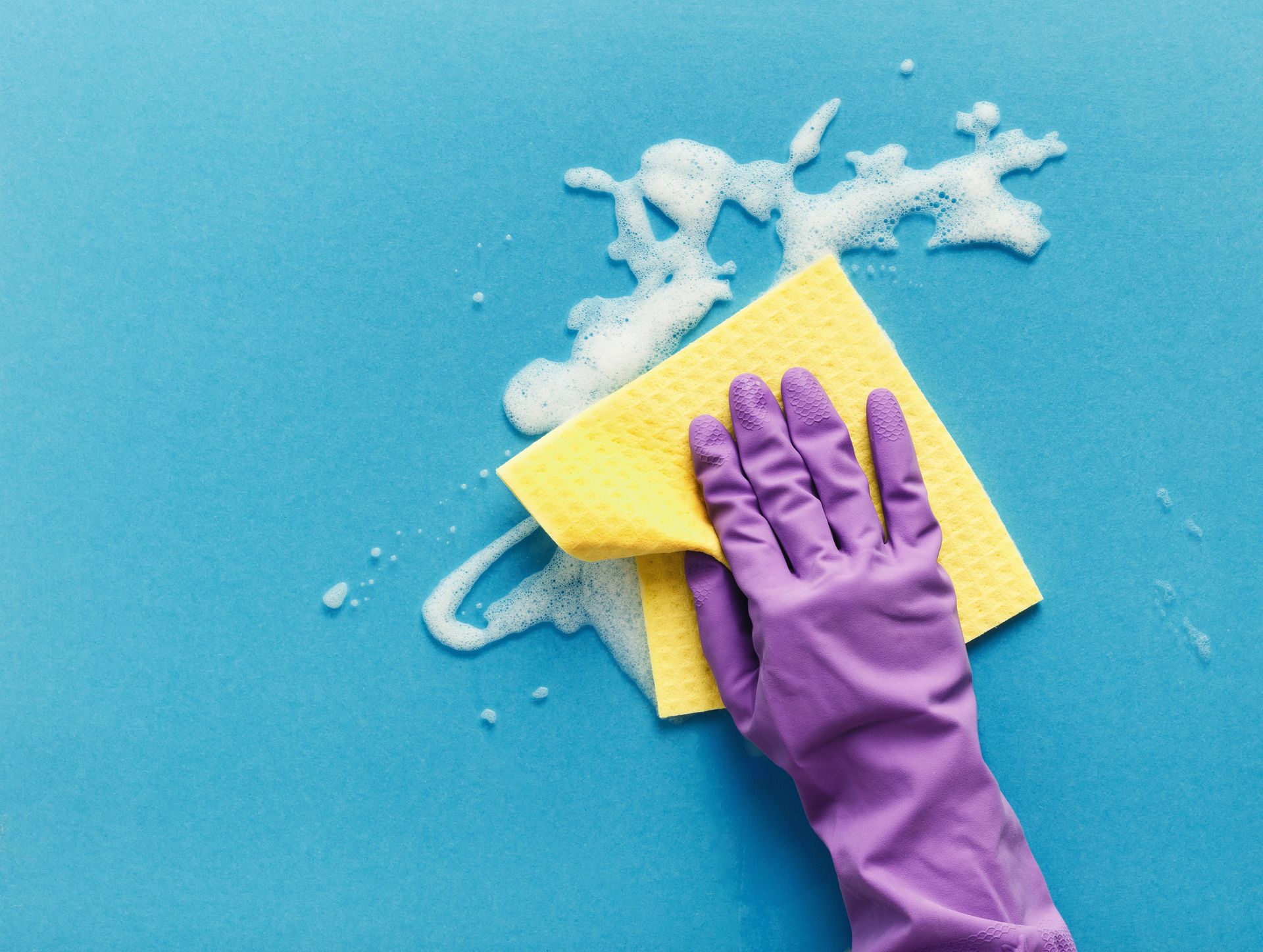 Mano guantata di viola che pulisce una superficie blu con una spugna gialla e schiuma di sapone.