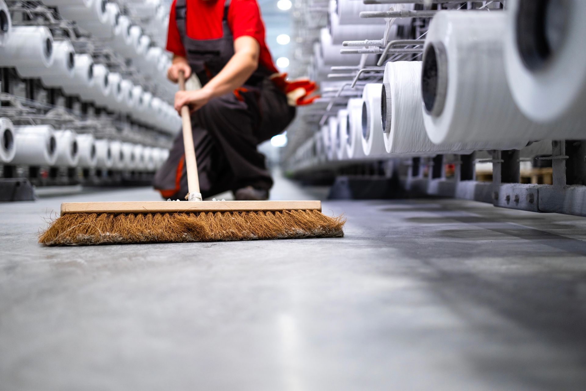 Un operaio spazza il pavimento nel corridoio di una fabbrica tessile, accanto a grandi macchine per la filatura.