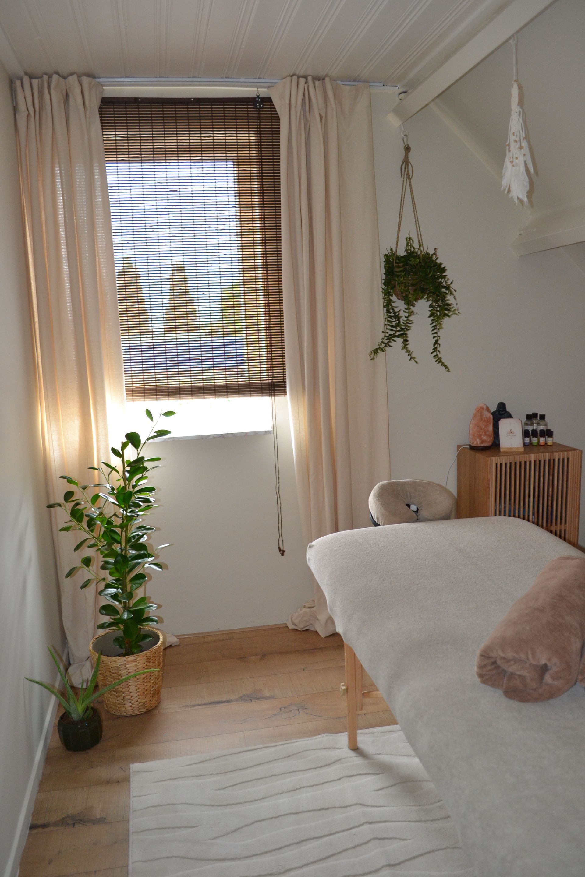 Gezellige massageruimte met wit bed, gestreept rolgordijn, crèmekleurige gordijnen, hangplanten en zacht natuurlijk licht.