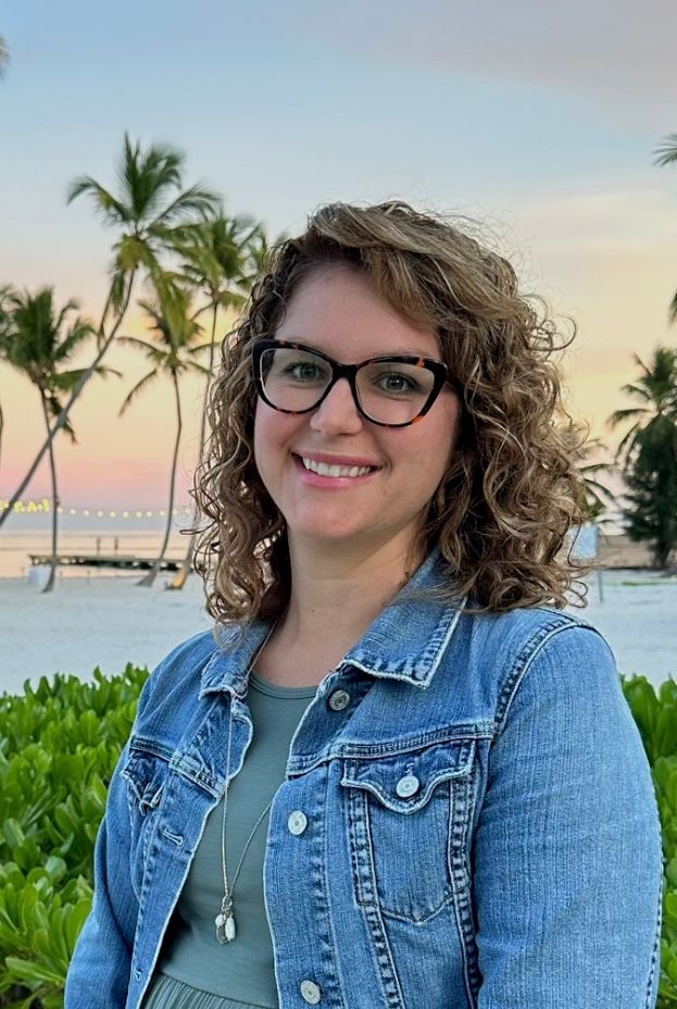 A woman wearing glasses and a denim jacket is smiling for the camera.