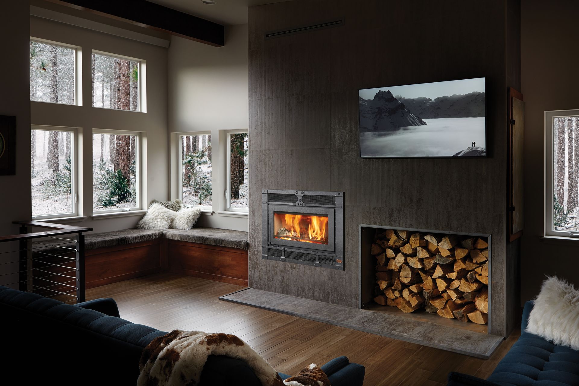 Cozy living room with a lit fireplace, stacked firewood, large windows, and a wall-mounted TV