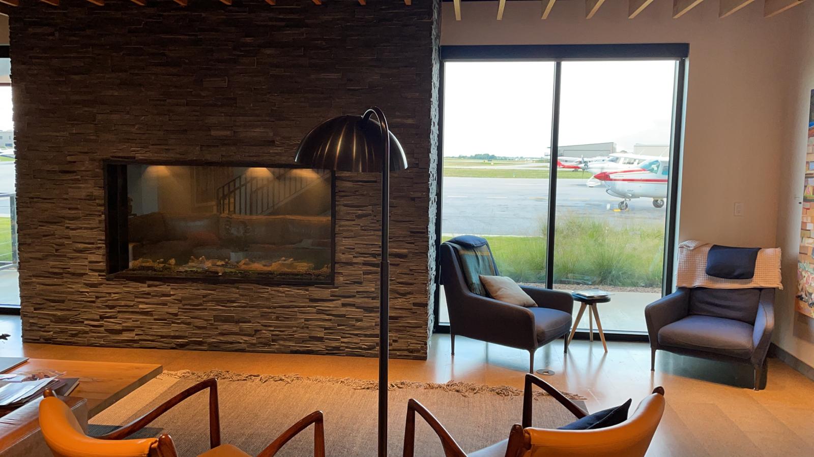 Modern lounge with stone fireplace, two armchairs, and large window overlooking an airport runway.