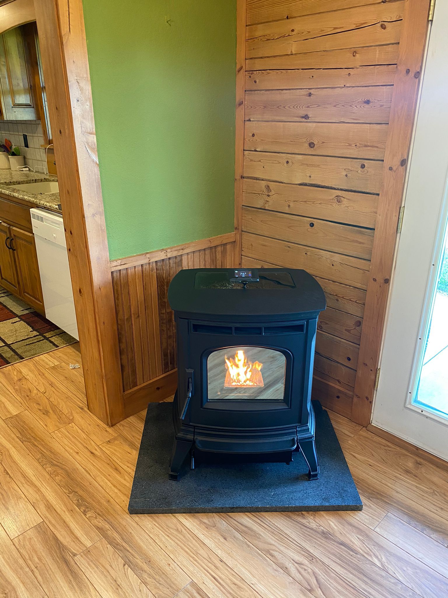 Black wood stove heating a cozy room with wood-paneled walls and hardwood floor.