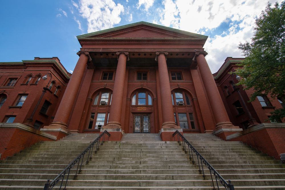 Red brick building with large columns, steps leading up to the entrance. Blue sky with clouds overhead.