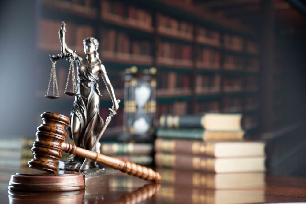 Gavel, statue of Lady Justice, and hourglass on a desk in a library, representing law and justice.