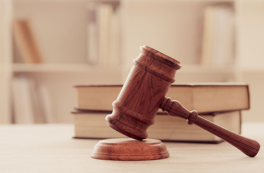 Wooden gavel on a stand, resting on a table with a stack of books, in front of a bookshelf.