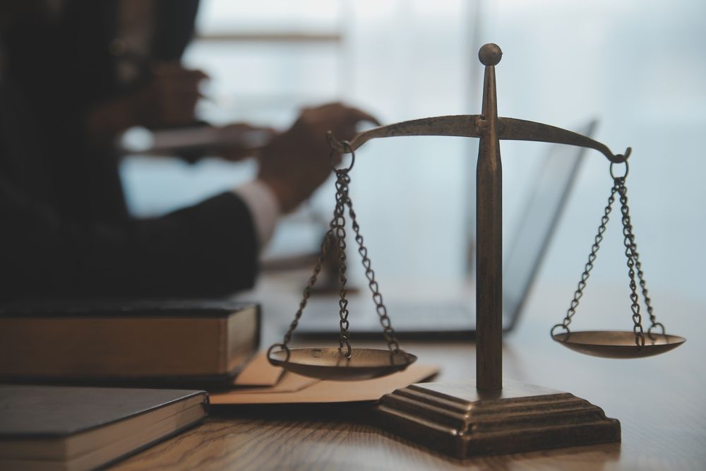 Scales of justice in focus, a person working on a laptop in background.