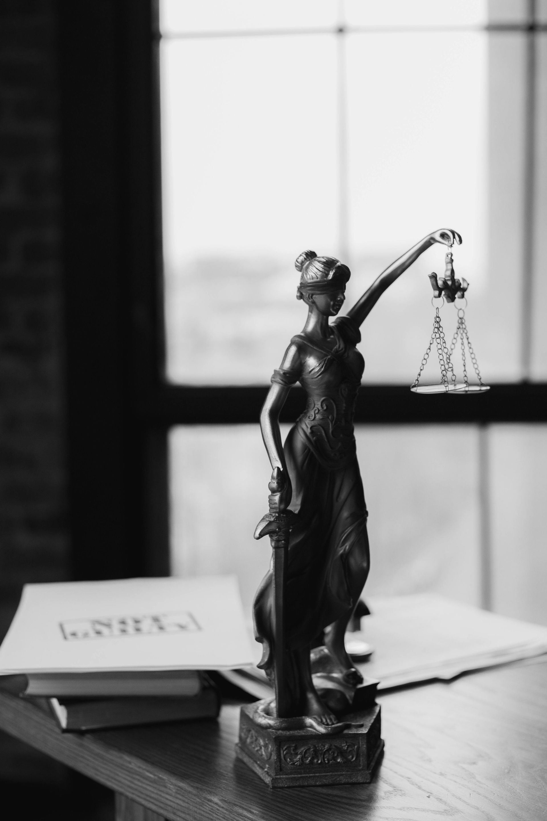 Statue of Lady Justice holding scales and sword, desk, and books in front of a window.