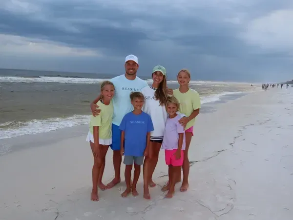 Family of six smiling on a beach under a cloudy sky.