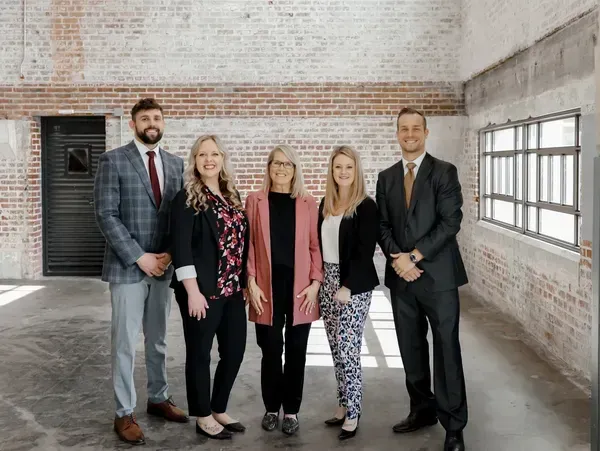 Five people, well-dressed, stand in a room with brick walls. They smile at the camera.