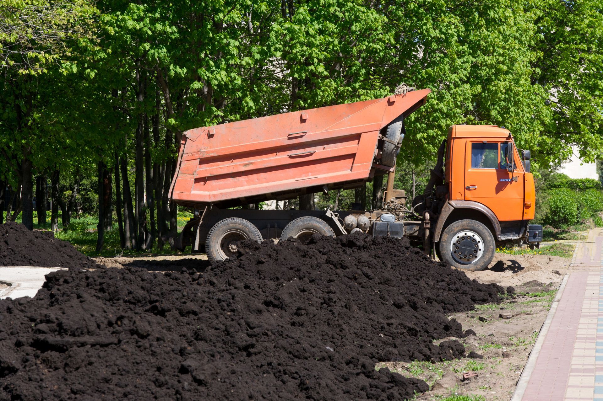Orange dump truck unloading soil beside a forested roadside