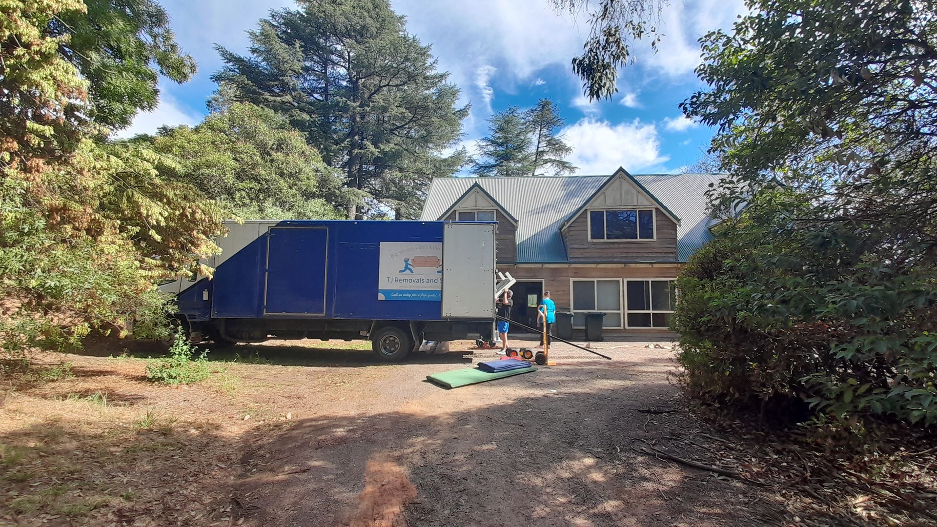 Blue moving truck parked in front of a house, with two people unloading items in a wooded driveway — TJ Removals and Storage in Wagga Wagga, NSW