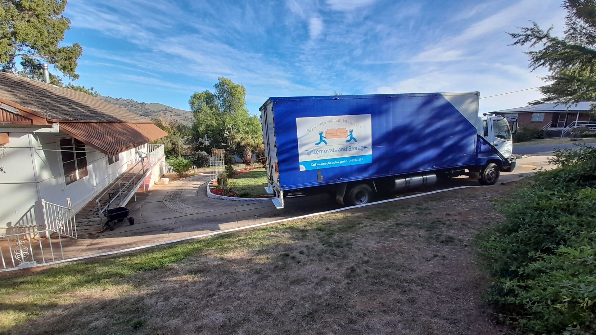 Blue delivery truck parked beside houses on a sunny dirt driveway, with trees and grass nearby — TJ Removals and Storage in Wagga Wagga, NSW