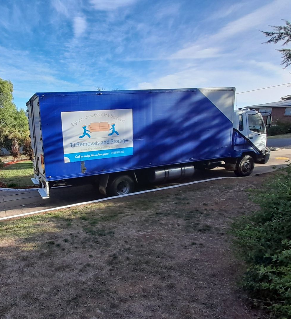 Blue delivery truck parked beside a house in a sunny yard, with trees and grass around it — TJ Removals and Storage in Wagga Wagga, NSW