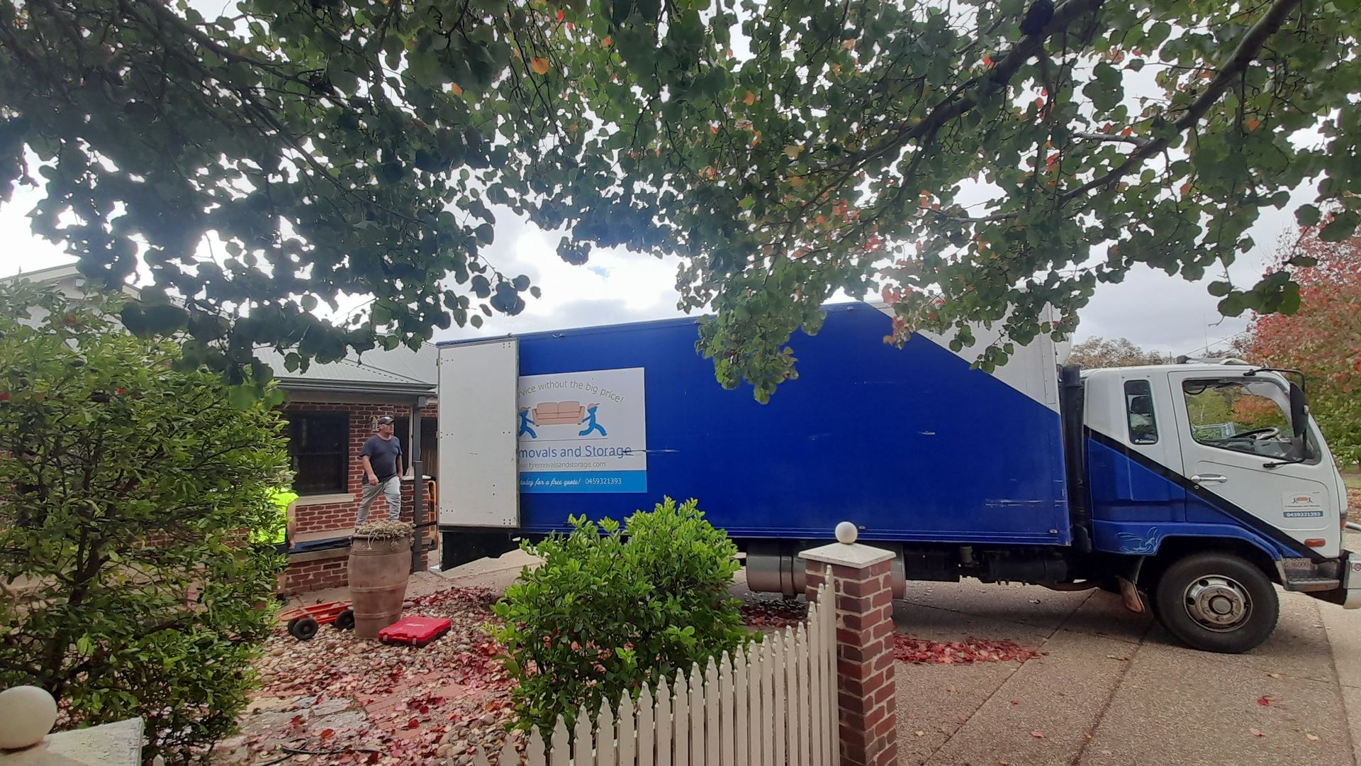 Blue delivery truck parked beside a house under trees with a white picket fence — TJ Removals and Storage in Wagga Wagga, NSW