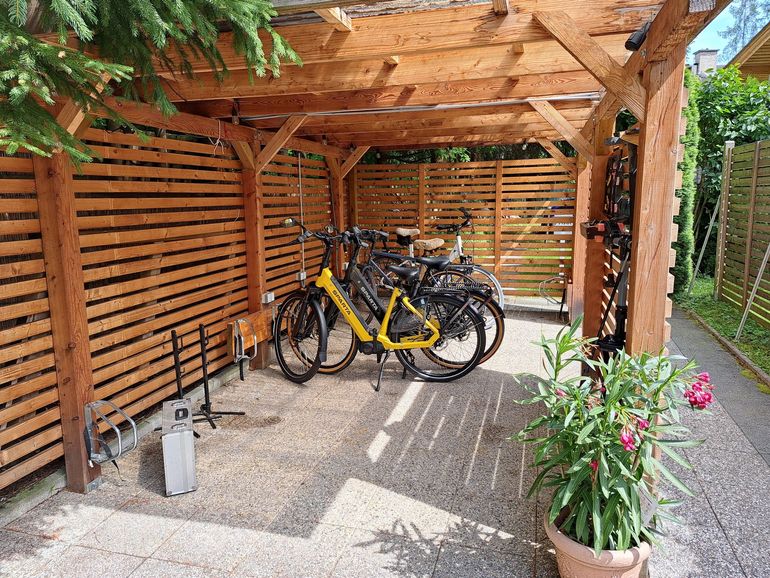 Überdachter Fahrradunterstand aus Holz mit mehreren geparkten Fahrrädern, darunter ein gelbes, neben Topfblumen.