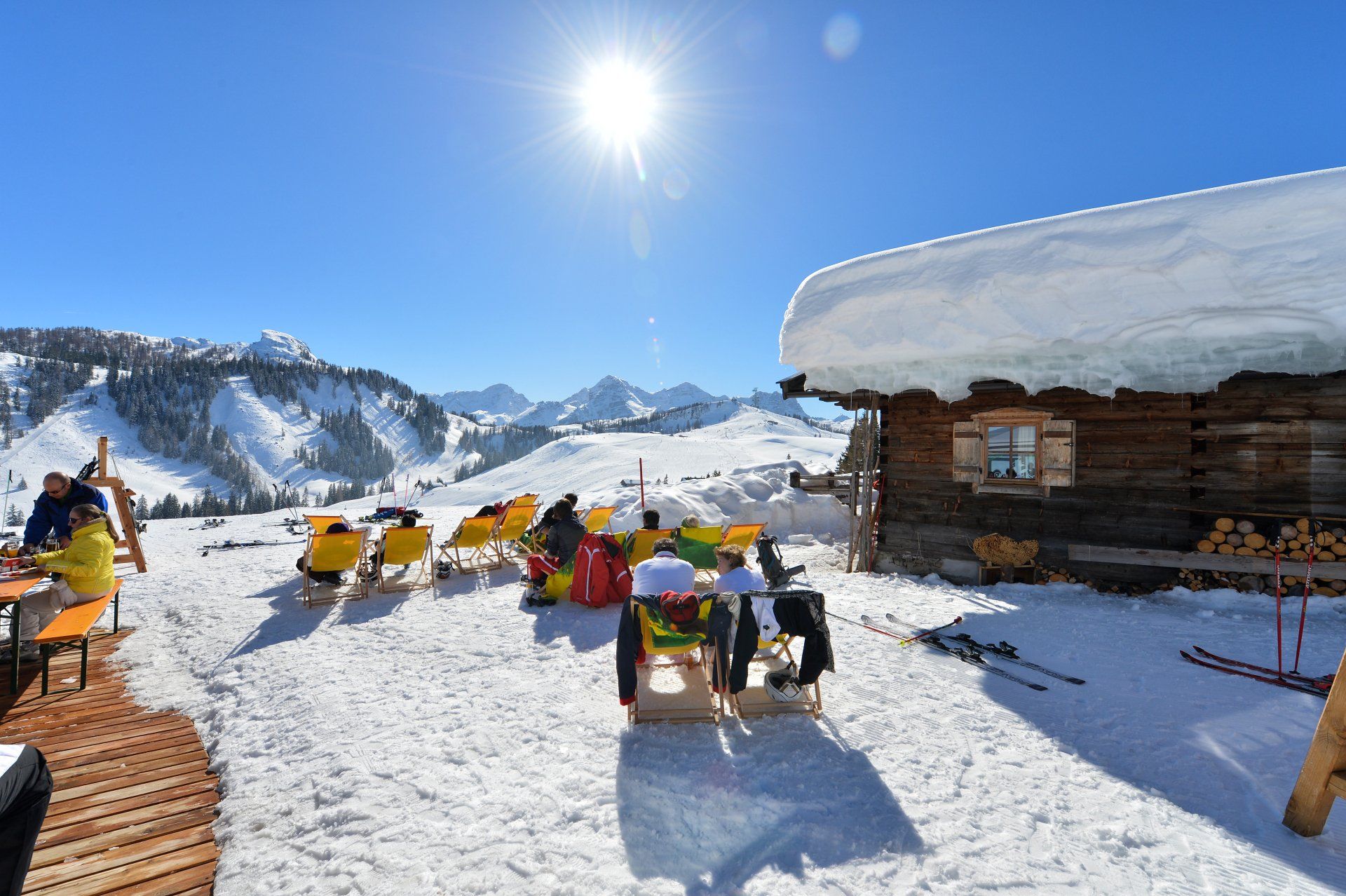 Terrasse einer Skihütte mit gelben Stühlen im strahlenden Schnee, Berge unter klarem, blauem Himmel.