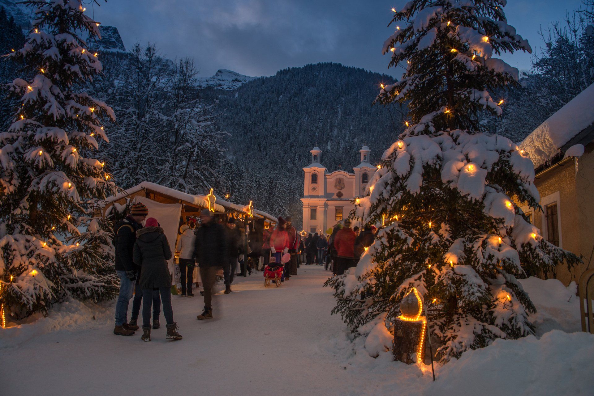 Verschneite Dorfstraße mit Menschen, die an beleuchteten Weihnachtsbäumen und erleuchteten Hütten vorbeigehen