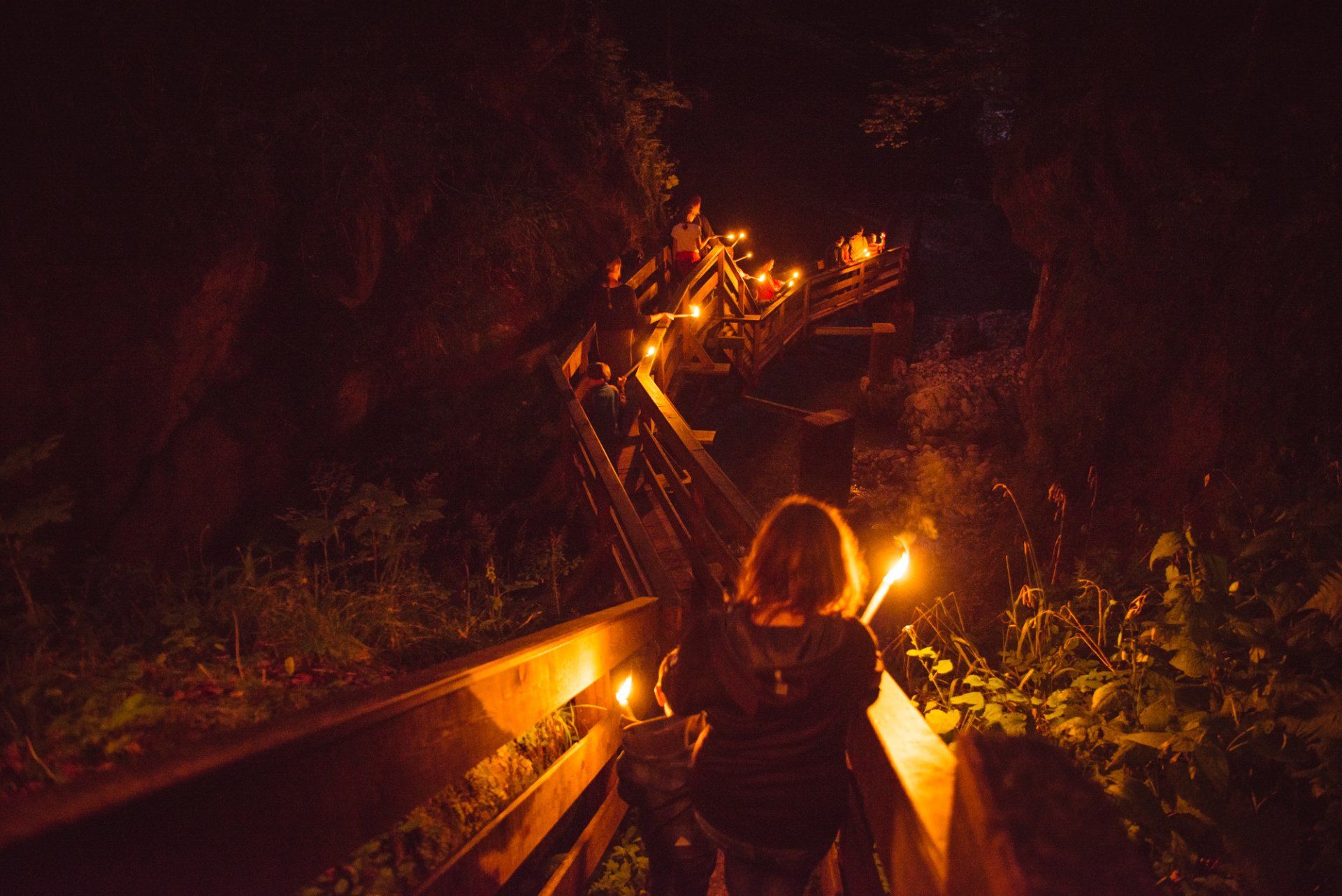 Eine Person auf einer Holzbrücke bei Nacht, beleuchtet von orangefarbenen Laternen in einem dunklen Wald