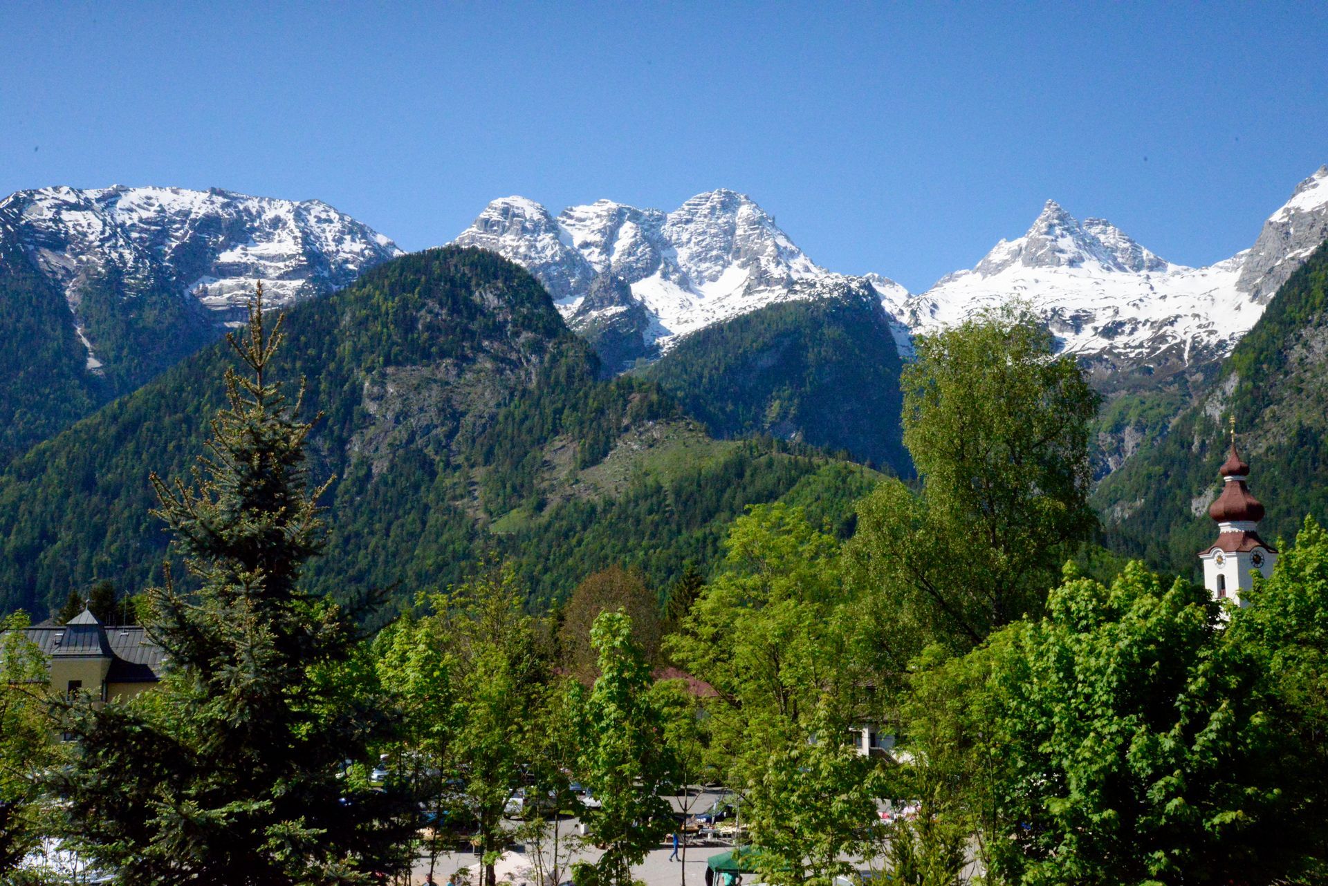 Schneebedeckte Berge über einem üppig grünen Wald mit einem kleinen Kirchturm unter blauem Himmel