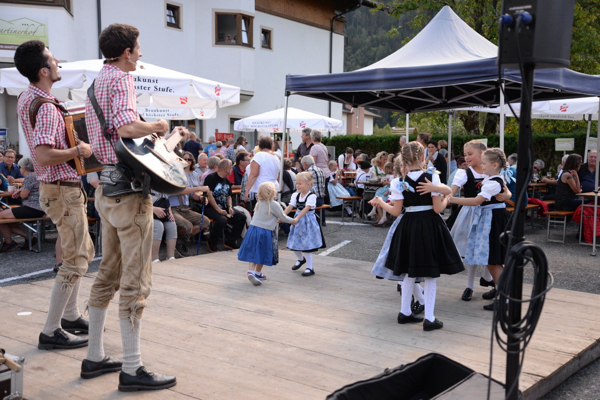 Musiker und Kinder treten bei einem Dorffest unter einem weißen Baldachin im Freien auf.