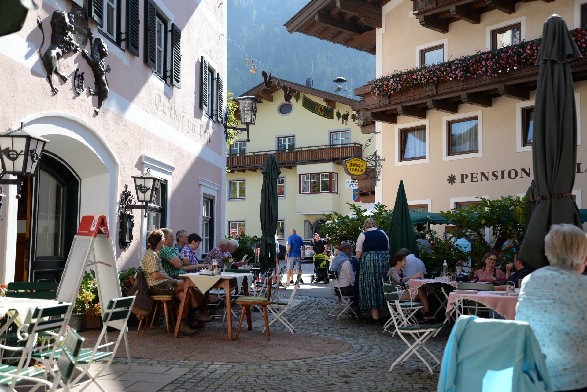 Café-Innenhof im Freien mit Menschen, die an Tischen neben weißen Gebäuden und Sonnenschirmen sitzen.