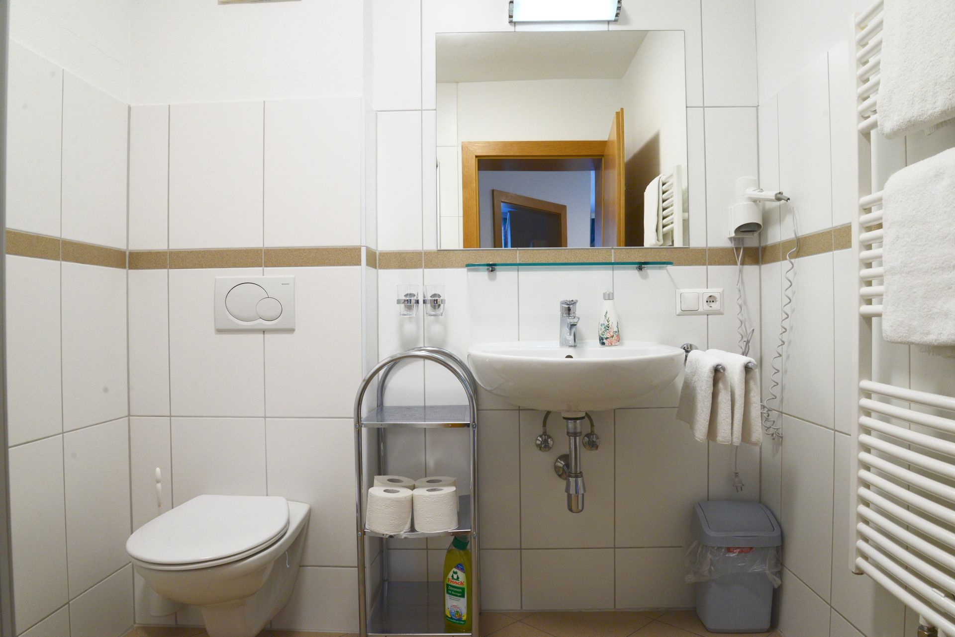 Helles, weiß gefliestes Badezimmer mit Toilette, Waschbecken, Spiegel, Handtuchhalter und kleinem Aufbewahrungswagen