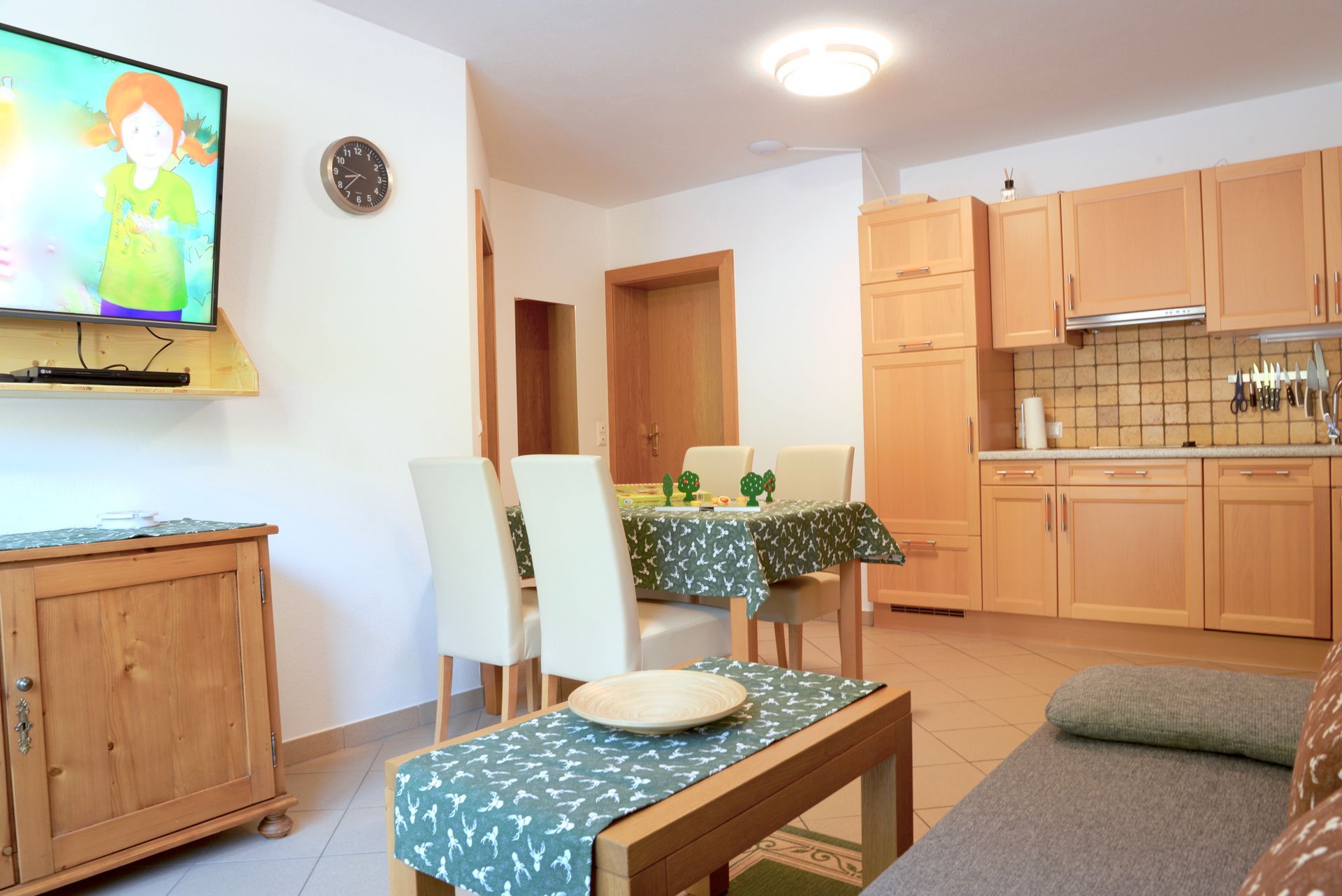 Helle Wohnküche mit Holzschränken, Tisch, Stühlen und einem an der Wand montierten Fernseher.
