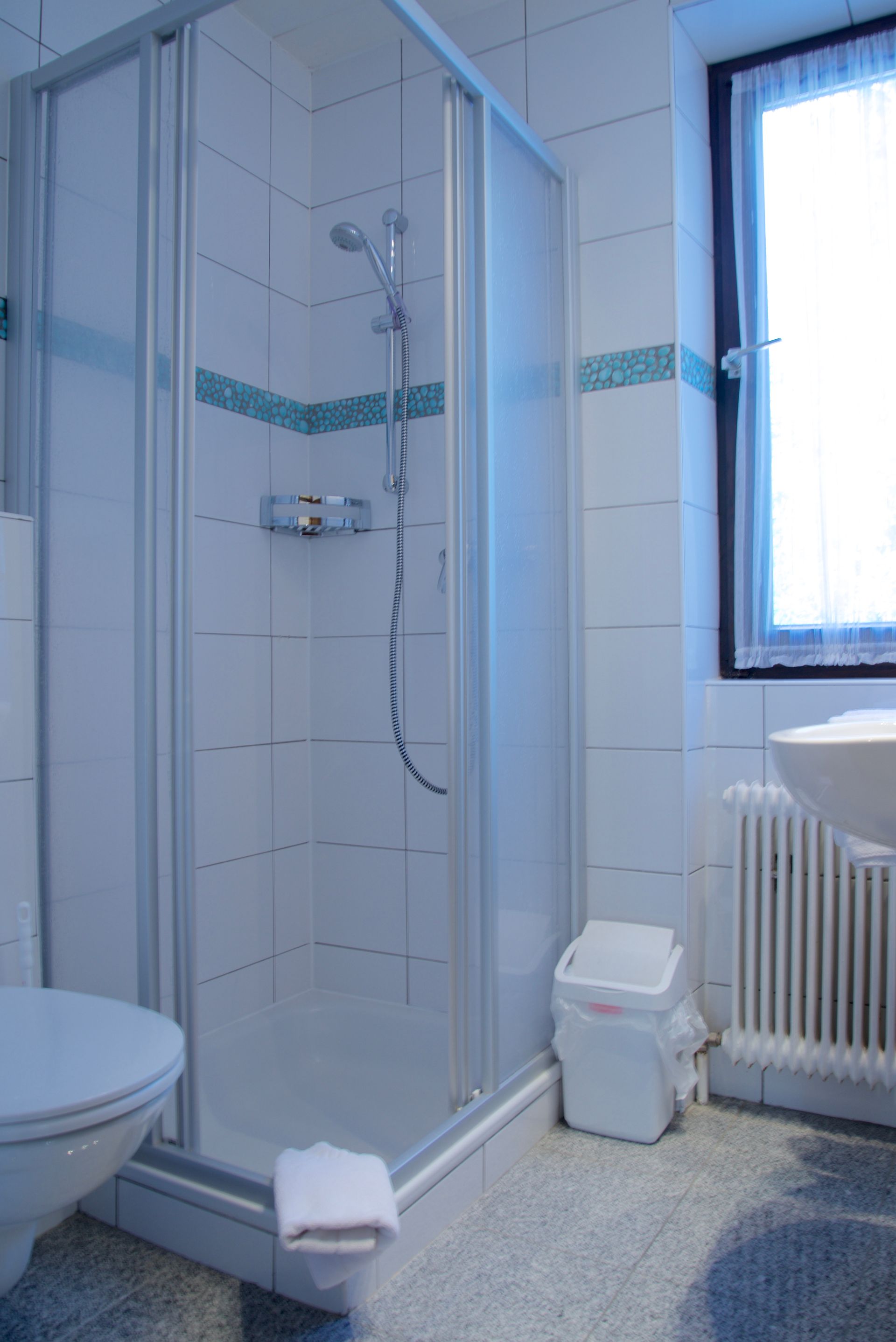 Kleines, gefliestes Badezimmer mit Eckdusche, WC, Fenster und Heizkörper